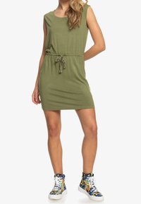 Olivově zelené šaty bez rukávů s tkaničkou v pase; hladká textura; v kombinaci s barevnými vzorovanými vysokými botami.