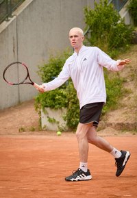 Muški sportaš u bijelom zip-up jakni, crnim šortsevima i crnim tenisicama, drži reket na terenu za tenis od gline s zelenilom u pozadini.