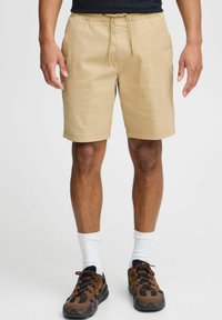 Khaki shorts with an elastic waistband and drawstring, featuring side pockets and a straight cut. Worn with brown and black sneakers.