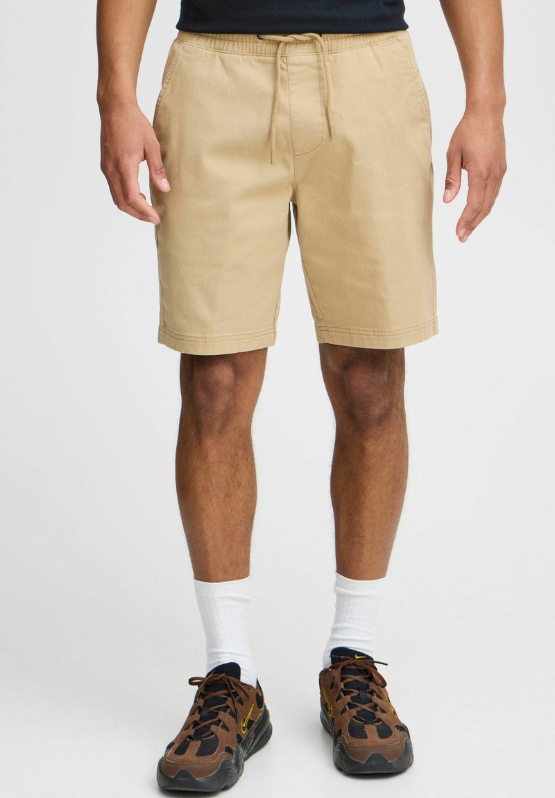 Khaki shorts with an elastic waistband and drawstring, featuring side pockets and a straight cut. Worn with brown and black sneakers.