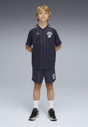 Niño rubio de pie con las manos detrás de la espalda, vistiendo un uniforme de fútbol con rayas negras y botas Puma negras sobre un fondo gris degradado.