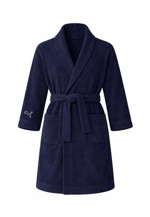 Frau mit langen blonden Haaren, die einen marineblauen Plüschbademantel mit Gürtel und vorderen Taschen trägt und mit der Hand in der Tasche lächelt.