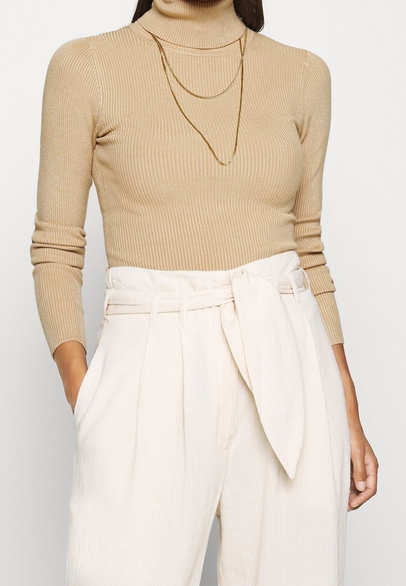 Femme portant un pull à col roulé beige côtelé, un pantalon blanc taille haute à jambes larges avec des plis, une ceinture et des colliers chaînes dorés superposés.