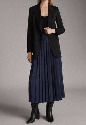 Woman wearing a black blazer, black top, navy pleated midi skirt, and black heeled ankle boots standing against a plain gray background.