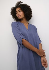 Robe légère bleue avec un col en V, des manches retroussées sécurisées par des boutons et une coupe décontractée, dotée d'une texture lisse et d'un design simple.