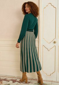 Top verde in maglia con maniche lunghe abbinato a una gonna a righe lungo caviglia in verde scuro e crema. Stivaletti beige con tacco completano l'outfit.