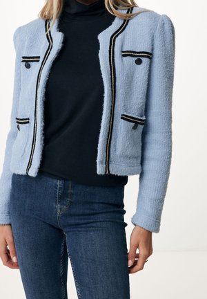 Vrouw die een lichtblauwe gebreide cardigan draagt met zwarte bies en knopen, een zwarte coltrui en donkerblauwe spijkerbroek, staand tegen een witte achtergrond.