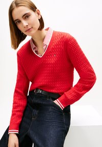Κόκκινο πλεκτό πουλόβερ με υφασμένο μοτίβο και λαιμόκοψη σε σχήμα V, με ριγέ μανσέτες, σε συνδυασμό με σκούρο μπλε τζιν και μαύρη ζώνη.