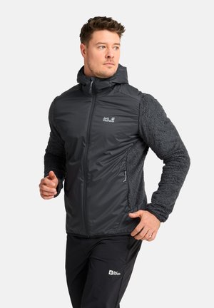 Mann trägt eine schwarz-graue Jack Wolfskin Kapuzenjacke und schwarze Hose und posiert mit leicht erhobenen Händen vor einfarbigem Hintergrund.