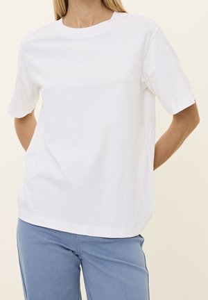Femme portant un t-shirt blanc uni à manches courtes et un pantalon bleu clair, debout les mains derrière le dos.
