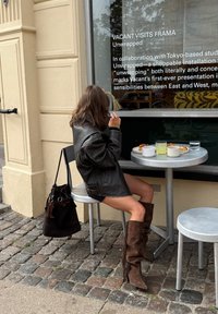 Chaqueta oversized de cuero negro, pantalones cortos de denim cortos, botas de ante marrón hasta la rodilla con flecos. Bebidas de plástico y cerámica sobre una mesa de metal.