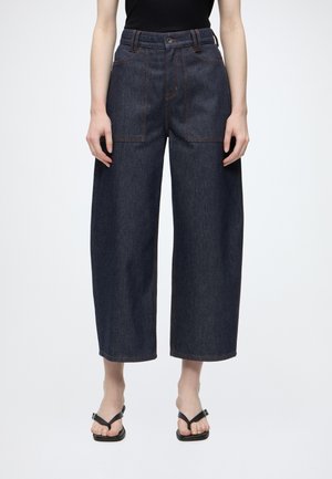 Personne portant un jean en denim foncé taille haute à jambes larges avec de grandes poches avant et des sandales noires à bout ouvert sur un fond uni.