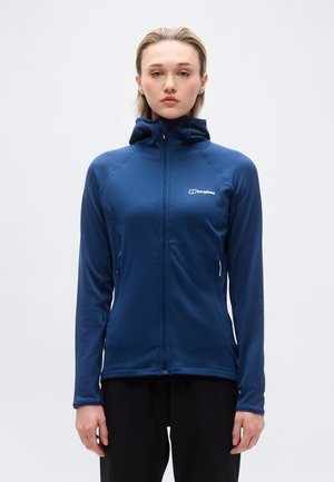 Noor naine seisab, kandes sinist kapuutsi ja tõmblukuga Berghausi jope, käed rahulikult kõrval, taustal hele puhas taust.