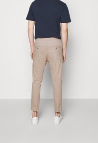 Beige chino's met een taps toelopende pasvorm, voorzien van een platte tailleband en twee achterzakken met metalen knopen, gecombineerd met witte sneakers.