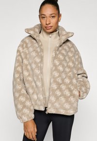 Ζακέτα από φλις σε μπεζ με διακριτικό σχέδιο, με ψηλό γιακά, κλείσιμο με φερμουάρ και ριμπ μανσέτες για άνετη εφαρμογή.