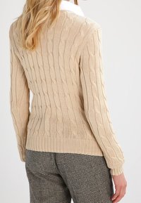 Pull en tricot câble beige avec ourlet et poignets côtelés. Le pull présente un motif texturé et est porté par-dessus un pantalon à carreaux.