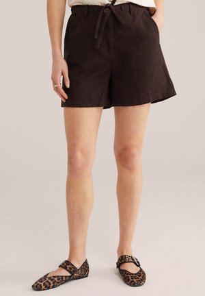 Frau trägt schwarze Shorts mit gebundenem Taillenband und Mary Jane Ballerinas mit Leopardenmuster, steht mit einer Hand in der Tasche vor hellem Hintergrund.