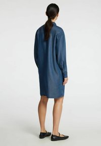 Tummansininen denim-paidan mekko, pitkät hihat, napitettu hihansuu ja rento mitoitus. Sileä kankaan rakenne ja suora helma. Mustat litteät kengät.