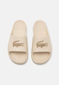 Sandale slide bej din cauciuc, cu o talpă texturată. Prezintă un logo de crocodil într-o nuanță de maro contrastant pe breteaua exterioară.
