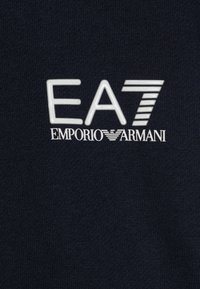 EA7 Emporio Armani-logotyp i vit tryck på en mörk marinblå stickad tygbakgrund.