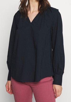 Kobieta w czarnej bluzce z długim rękawem, dekoltem w kształcie litery V i marszczonymi mankietami oraz różowych spodniach, stojąca na jednolitym jasnym tle.