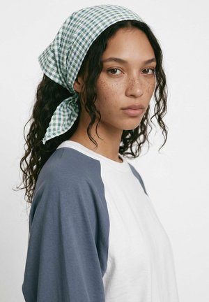 Une femme aux longs cheveux foncés porte un foulard à carreaux verts et blancs et une chemise blanche à manches bleu-gris, sur un fond uni.
