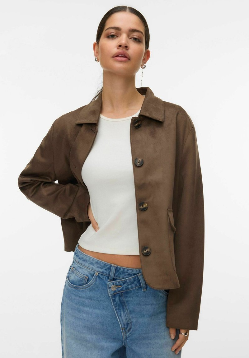 Veste en daim marron avec fermeture à boutons, col pointu et poches latérales, portée avec un haut blanc et un jean en denim bleu.