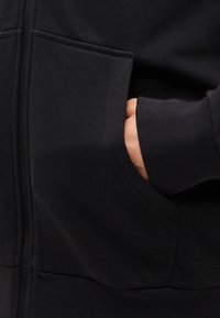 Χέρι μερικώς μέσα στην μπροστινή τσέπη μιας μαύρης ζακέτας με φερμουάρ, με εμφανές ριμπ μανσέτα στο μανίκι.