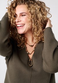 Olijfgroene trui met een V-halsdesign en brede mouwen, gecombineerd met een gelaagde, meerstrengige ketting met een textuur en aardetinten en vormen.