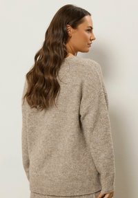 Beżowy sweter dzianinowy o luźnym kroju, z okrągłym dekoltem, prążkowanymi mankietami oraz miękkim, teksturowanym wykończeniem. Bez widocznych wzorów.