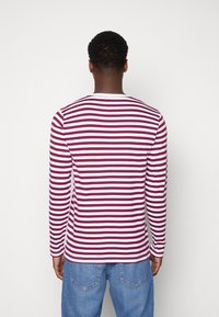Mann, der mit dem Rücken zur Kamera steht und ein langärmeliges weißes und dunkelrotes gestreiftes Hemd sowie blaue Jeans trägt, vor einem einfachen weißen Hintergrund.