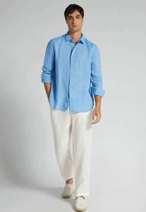 Mężczyzna stojący w jasno niebieskiej, długorękawowej koszuli zapinanej na guziki, białych luźnych spodniach i białych wsuwanych butach na jednolitym tle.