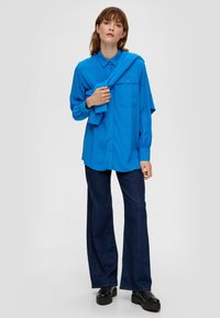 Blauwe overhemdd met een kraag en een zak, gecombineerd met donkere wijde jeans en zwarte plateau schoenen. Trui over de schouders gehangen.