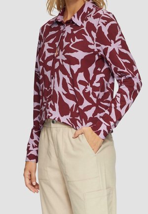 Nainen, jolla on päällään pitkähihainen nappipaita, jossa on viininpunaisia ja lilahtavia abstrakteja lehtikuvioita. Yhdistettynä on beigejä, löysästi istuvia housuja, joissa on sivutaskut.