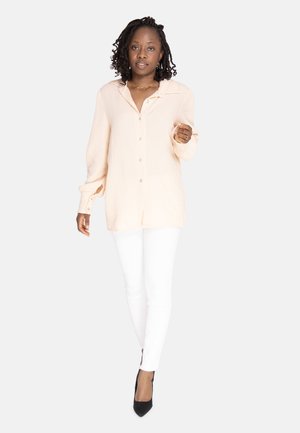 Camicia button-down rosa chiaro con tessuto texturizzato e colletto, abbinata a pantaloni bianchi aderenti e scarpe con tacco a punta nere.