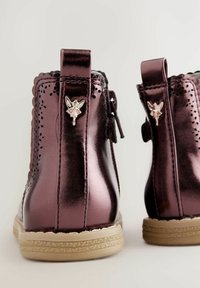 Fialové metalické kotníkové boty s vroubkovanými okraji, zdobené kouzlem víly a texturovanou béžovou podrážkou. Zapínání na zip pro snadné obouvání.