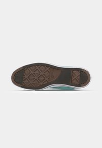 Semelle d’une basket basse turquoise montrant une semelle en caoutchouc texturée marron avec motif losange et logo de la marque sur fond blanc.