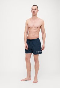 Sötétkék fürdőnadrág rugalmas derékpánttal és húzózsinórral, fehér EA7 Emporio Armani logóval a bal combon.