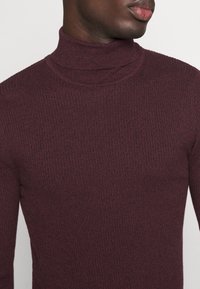 Pull à col roulé bordeaux avec une texture côtelée, design ajusté et col haut, présentant des variations subtiles de couleur et de motif.