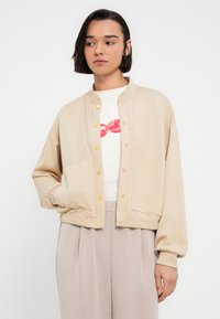 Μπεζ μπουφάν τύπου μπομπέρ με χρυσά κουμπιά πατιλέτα, ριμπ μανσέτες και δύο μπροστινές τσέπες. Φορεμένο πάνω από λευκό τ-shirt με γραφικό σχέδιο. Ομαλή υφή υφάσματος.