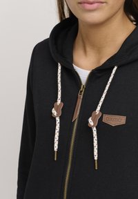 Svart zip-up hoodie med en mjuk textur, kontrasterande vita och flerfärgade dragsnören, samt en läderlogga på övre vänstra sidan.