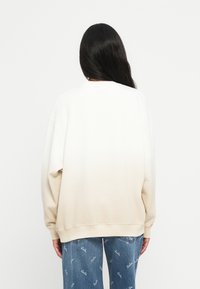 Sweatshirt oversized em creme, desvanecendo para um bege claro. Material de algodão macio com punhos e barra canelados, combinado com jeans azuis com padrão.