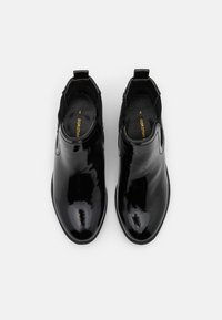 Para błyszczących czarnych lakierowanych botków za kostkę typu slip-on z uchwytami, widziana z góry na białym tle.