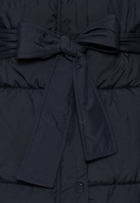 Navy blauwe pufferjack met een gestrikte stoffen riem, voorzien van een gewatteerde textuur en een drukknoopsluiting aan de voorkant.