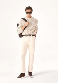 Suéter de punto color crema con diseño de trenzas, combinado con pantalones de un color claro. Los accesorios incluyen gafas de sol oscuras y una mochila marrón. Zapatillas marrones.