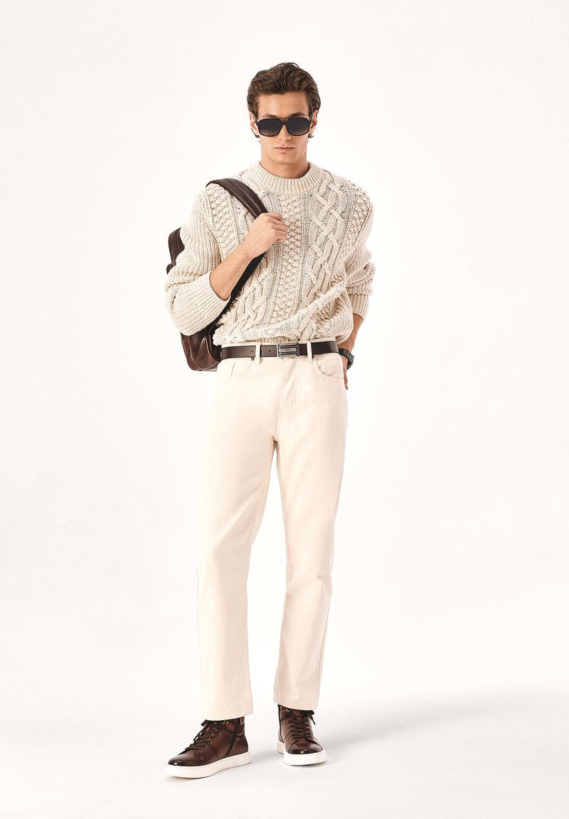 Suéter de punto color crema con diseño de trenzas, combinado con pantalones de un color claro. Los accesorios incluyen gafas de sol oscuras y una mochila marrón. Zapatillas marrones.