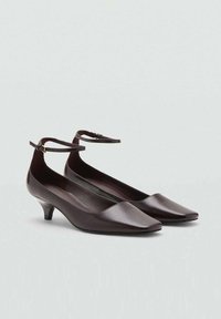 Zapatos bajos de cuero marrón oscuro con puntera puntiaguda y tiras delgadas en el tobillo con pequeñas hebillas doradas, exhibidos sobre un fondo claro y liso.