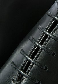 Chaussure en cuir noir avec des coutures visibles et des lacets. Texture lisse avec une finition brillante. Comprend cinq paires d'œillets pour le laçage.