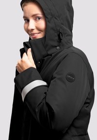 Chaqueta aislante negra con capucha, que presenta una franja plateada en la manga y un parche con logo en el hombro. Textura suave, cierre de cremallera.