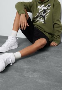Sudadera verde oliva con estampado gráfico blanco, pantalones cortos negros y zapatillas altas blancas con cordones texturizados y correas en el tobillo.